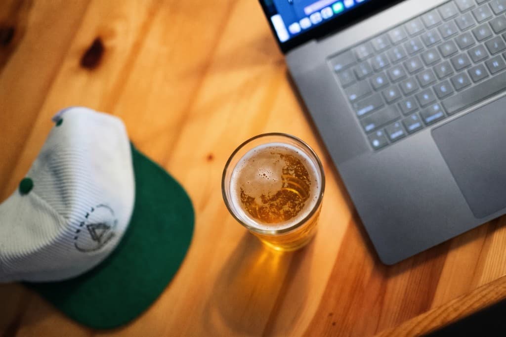 A pint of beer on a desk beside a laptop and a cap